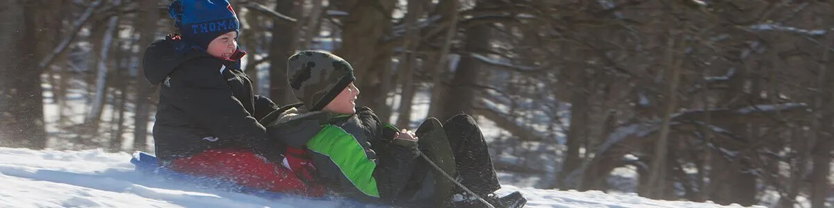 Two children are sledding down a snow covered hill.